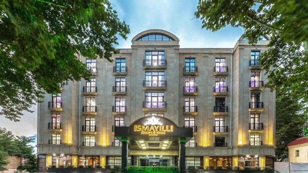 Un hotel de varios pisos con una entrada destacada, balcones y un jardín frontal arbolado.