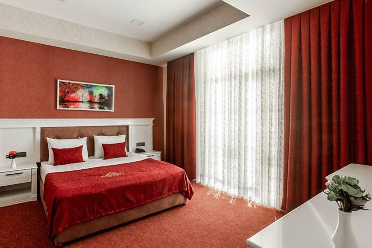 Una habitación de hotel con una cama grande, una mesita de noche blanca, una mesilla auxiliar, una manta roja, un cuadro en la pared y una ventana con cortinas.