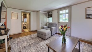 A spacious living room with a grey sofa, ottoman, and a wooden coffee table. There is a patterned rug on the floor, a door leading to another room, and a framed picture on the wall. - Highville Hideaway - 2 bed, 1 bath, country views (Conestoga)
