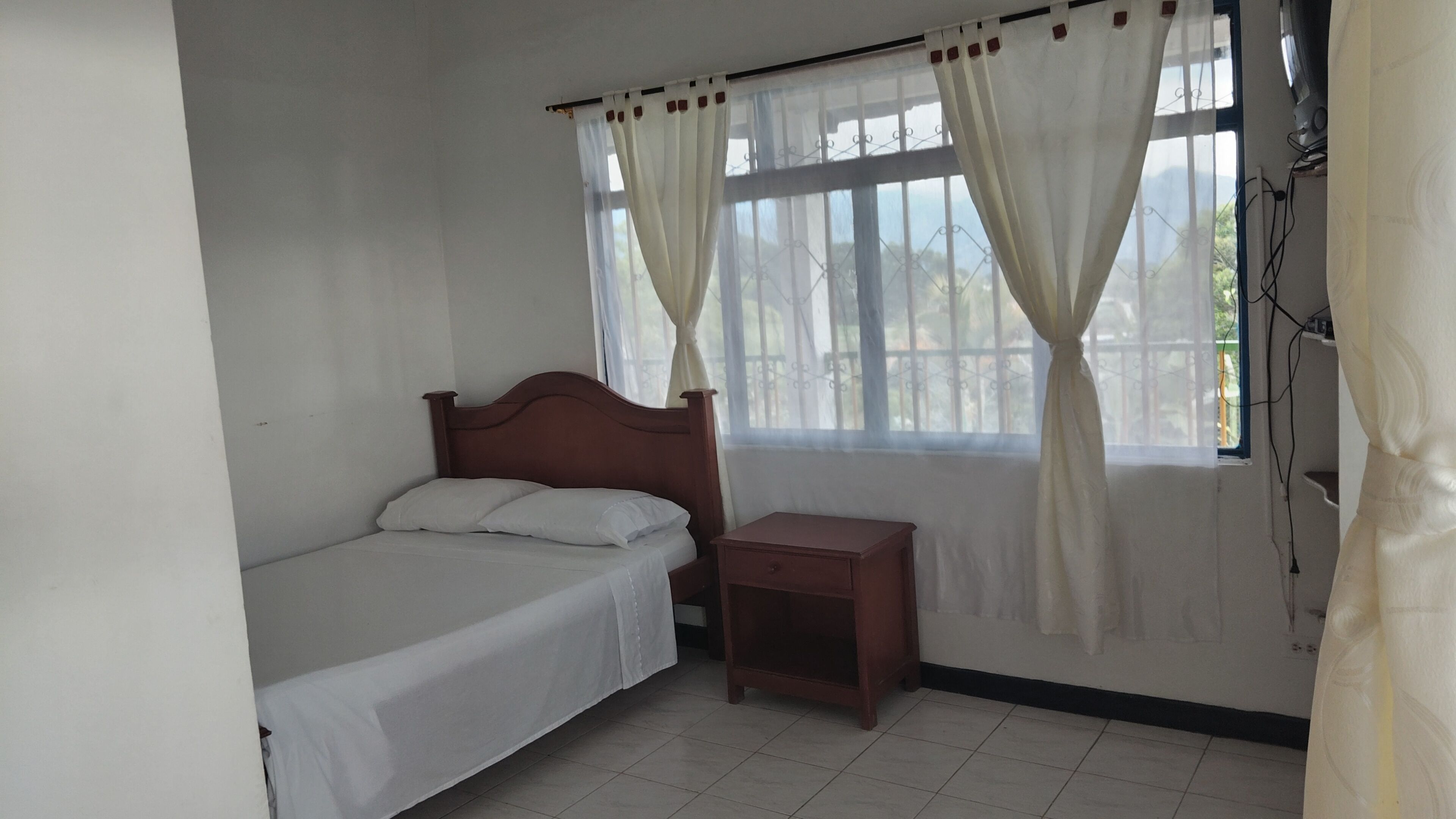 Uma cama de solteiro com cabeceira de madeira, uma mesinha de cabeceira e uma janela com cortinas.