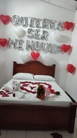 Una cama con un ramo, pétalos de rosa y globos plateados que deletrean un mensaje.