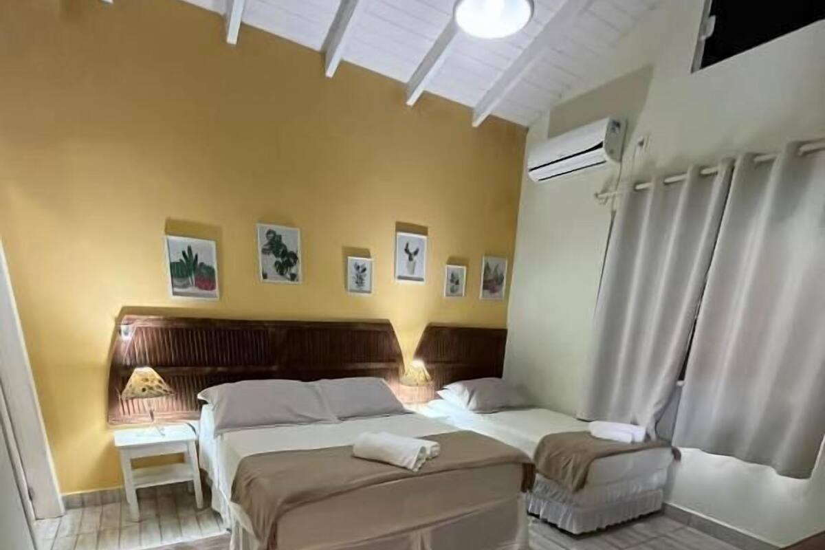 A hotel room with two beds, a wooden headboard, a nightstand, a lamp, a window with curtains, and a wall-mounted air conditioner.