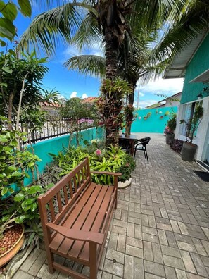 A wooden bench in a tropical garden setting with a tiled patio, a palm tree, and a view of a building with a blue wall and a fence. - Pousada Gengibre (Barra Velha)