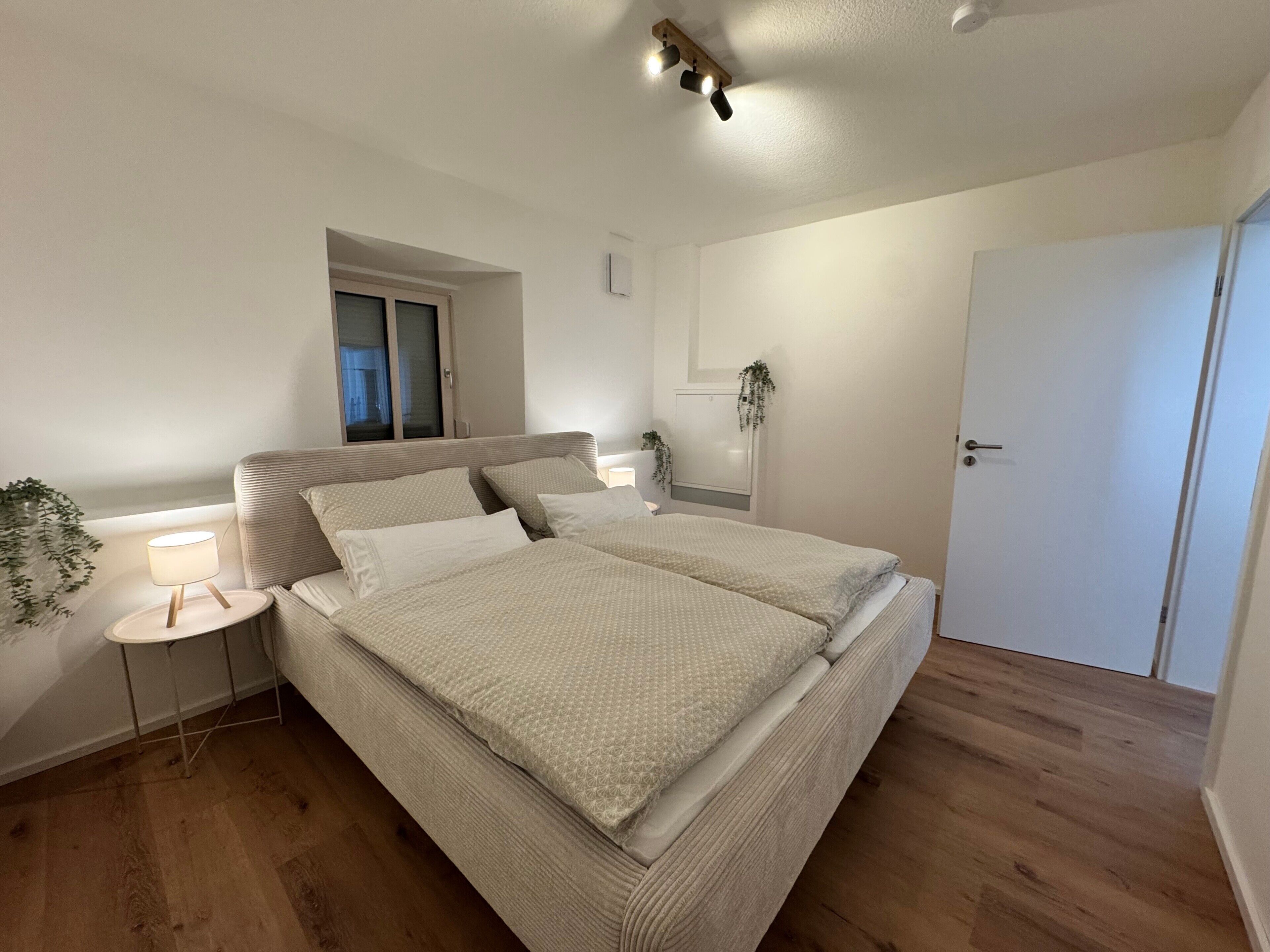 Ein Schlafzimmer mit Bett, Nachttischen, einem Fenster, einer Tür und einer Deckenleuchte.