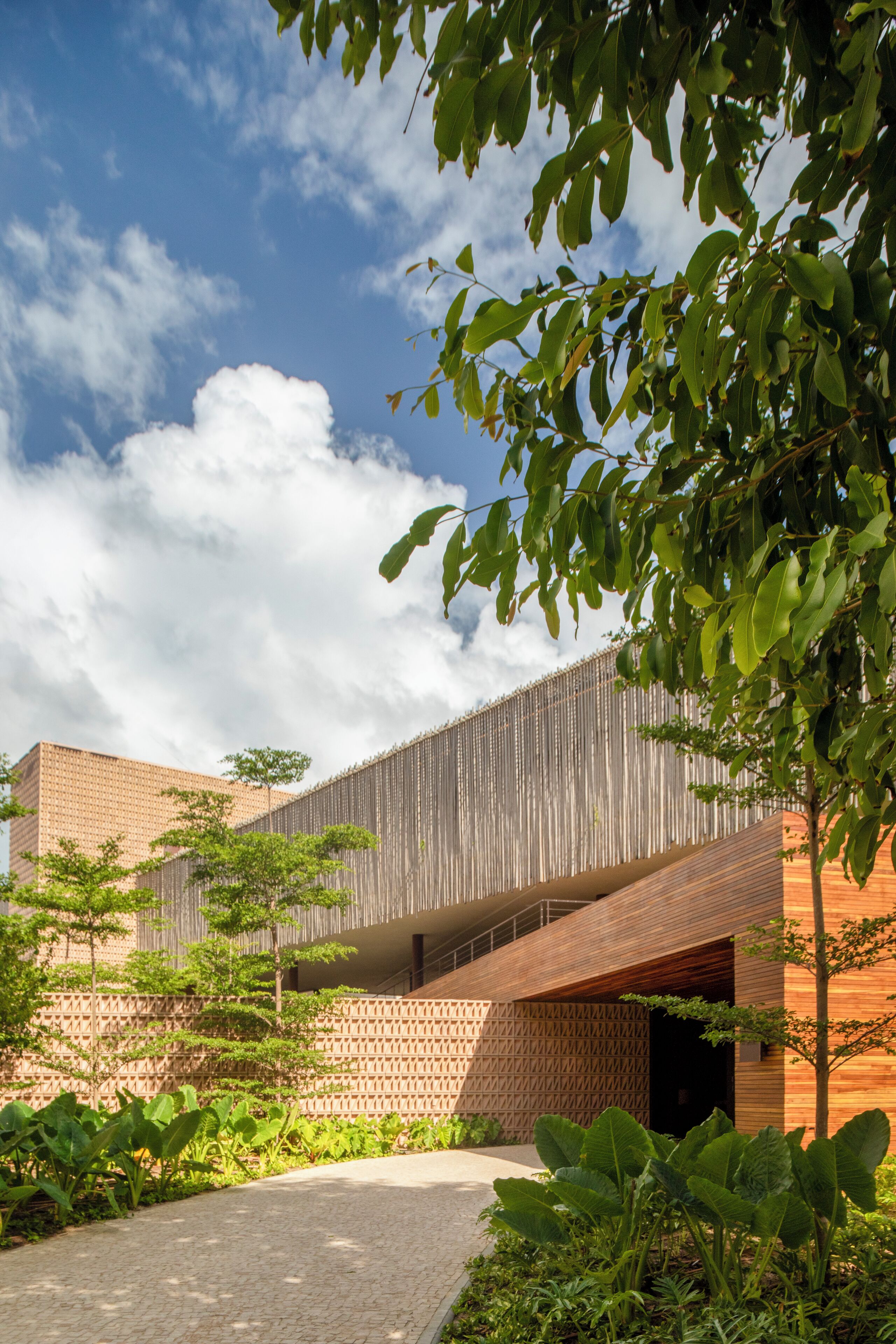 A modern building with a wooden facade, a brick wall, and a paved pathway surrounded by greenery.