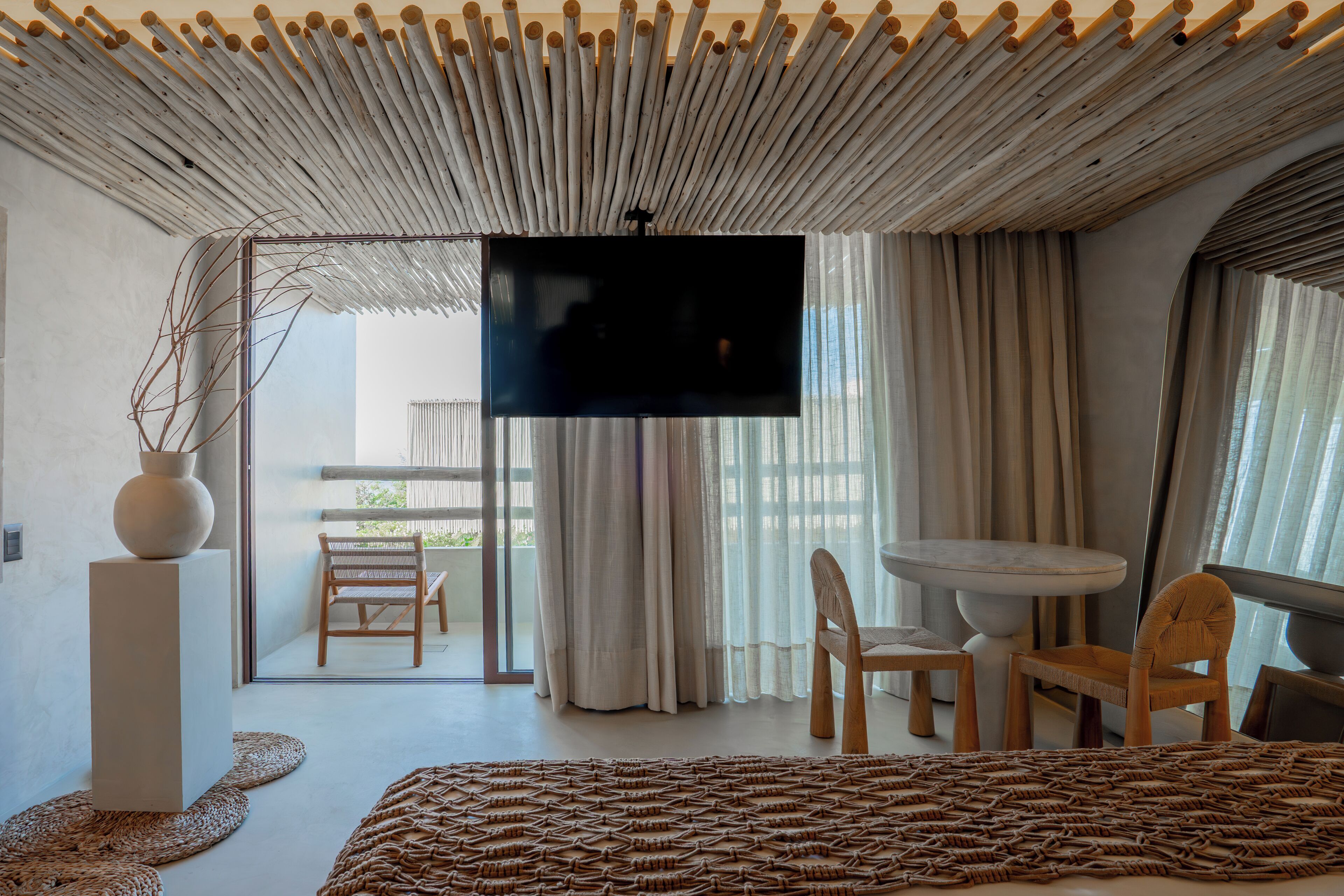 A modern hotel room with a large flat-screen TV, a balcony with a view, and a dining area with a round table and chairs.