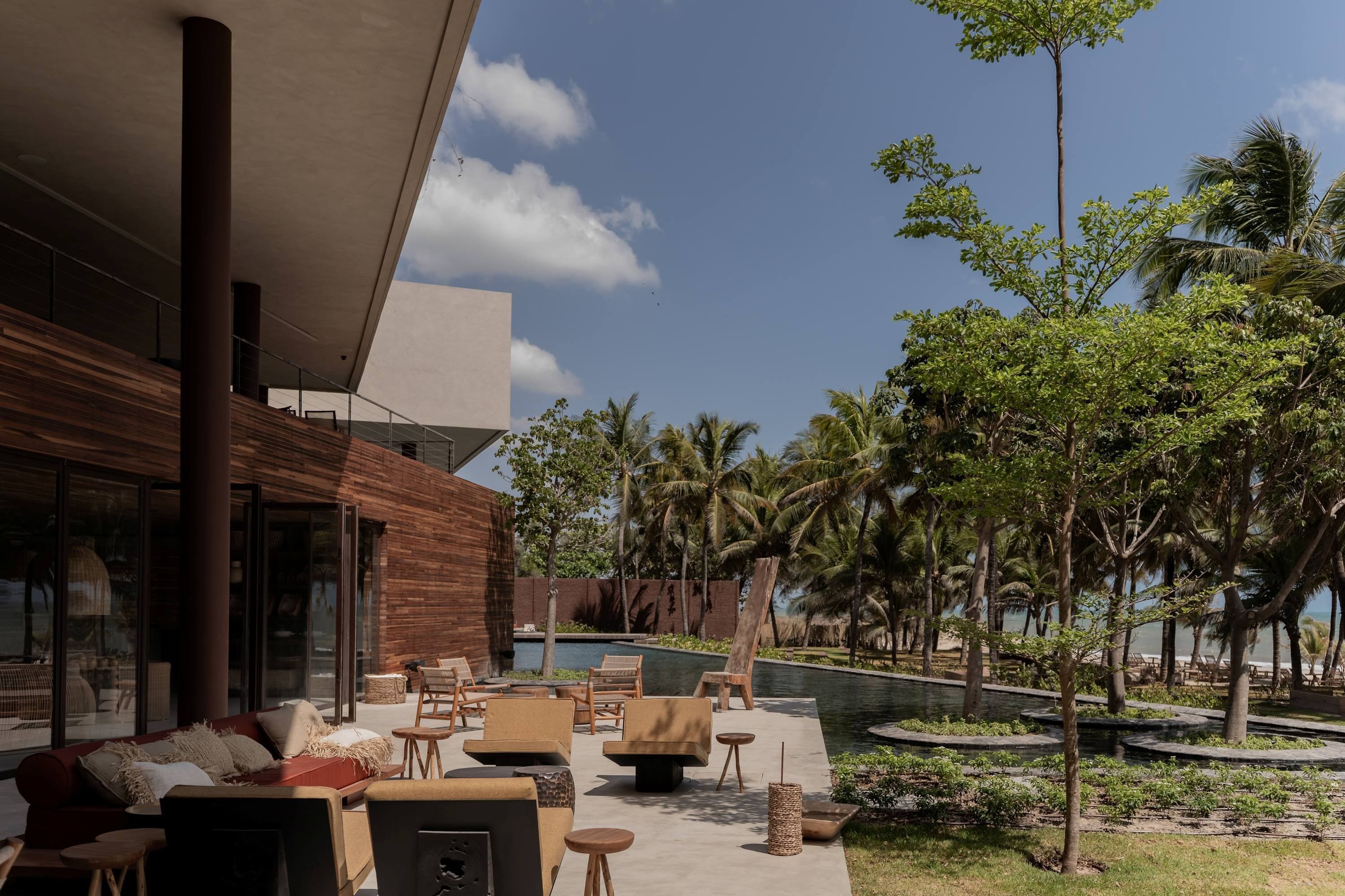 Uma área de estar ao ar livre moderna, com assentos e piscina, rodeada por palmeiras e vegetação.