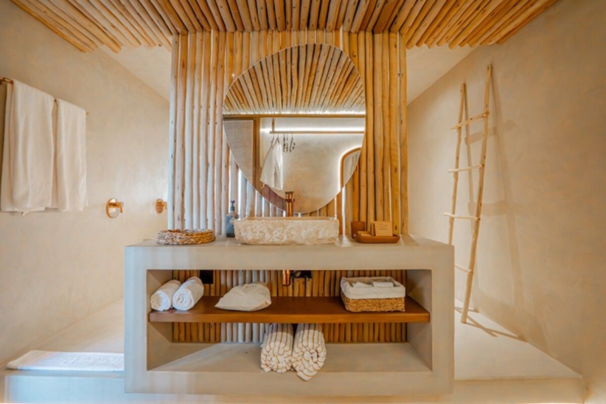 Une salle de bain avec un plafond en lambris de bois, un grand miroir, un lavabo et des étagères où sont rangées des serviettes et des articles de toilette.