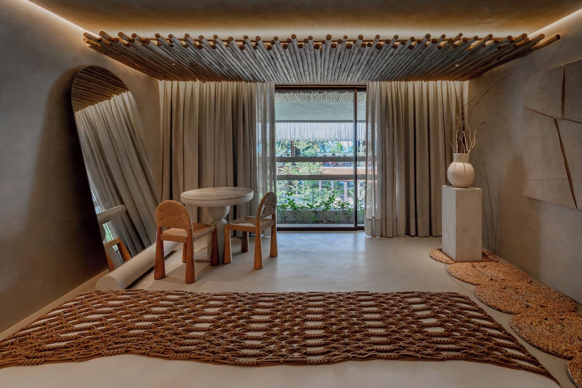 A modern bedroom with a large bed, a round table, and chairs, a view of greenery outside, and a unique ceiling design.
