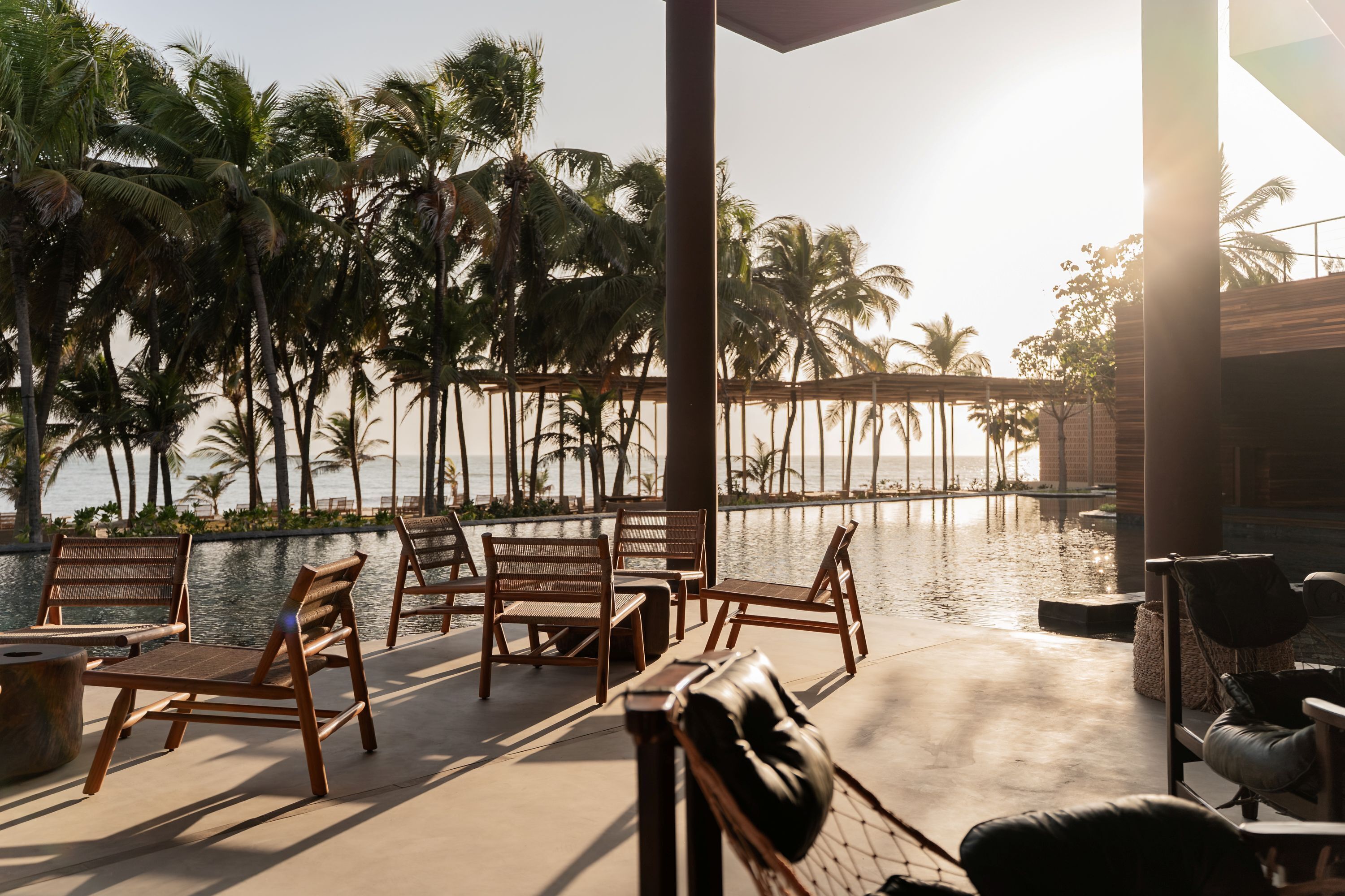 A sunlit outdoor seating area with wooden chairs and a view of palm trees and a body of water.