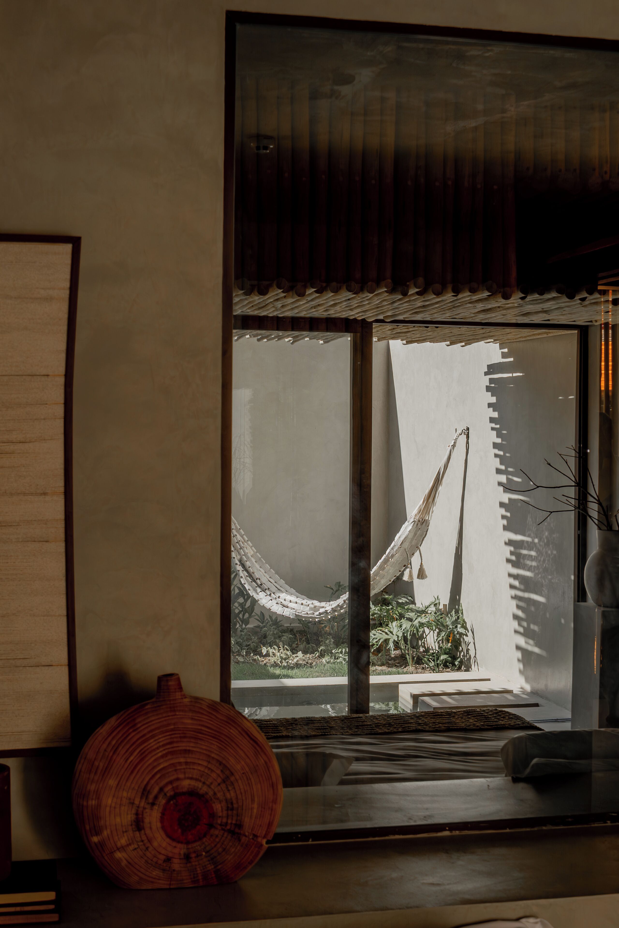 Una habitación con un ventanal grande, un florero de madera y vistas a una hamaca afuera.