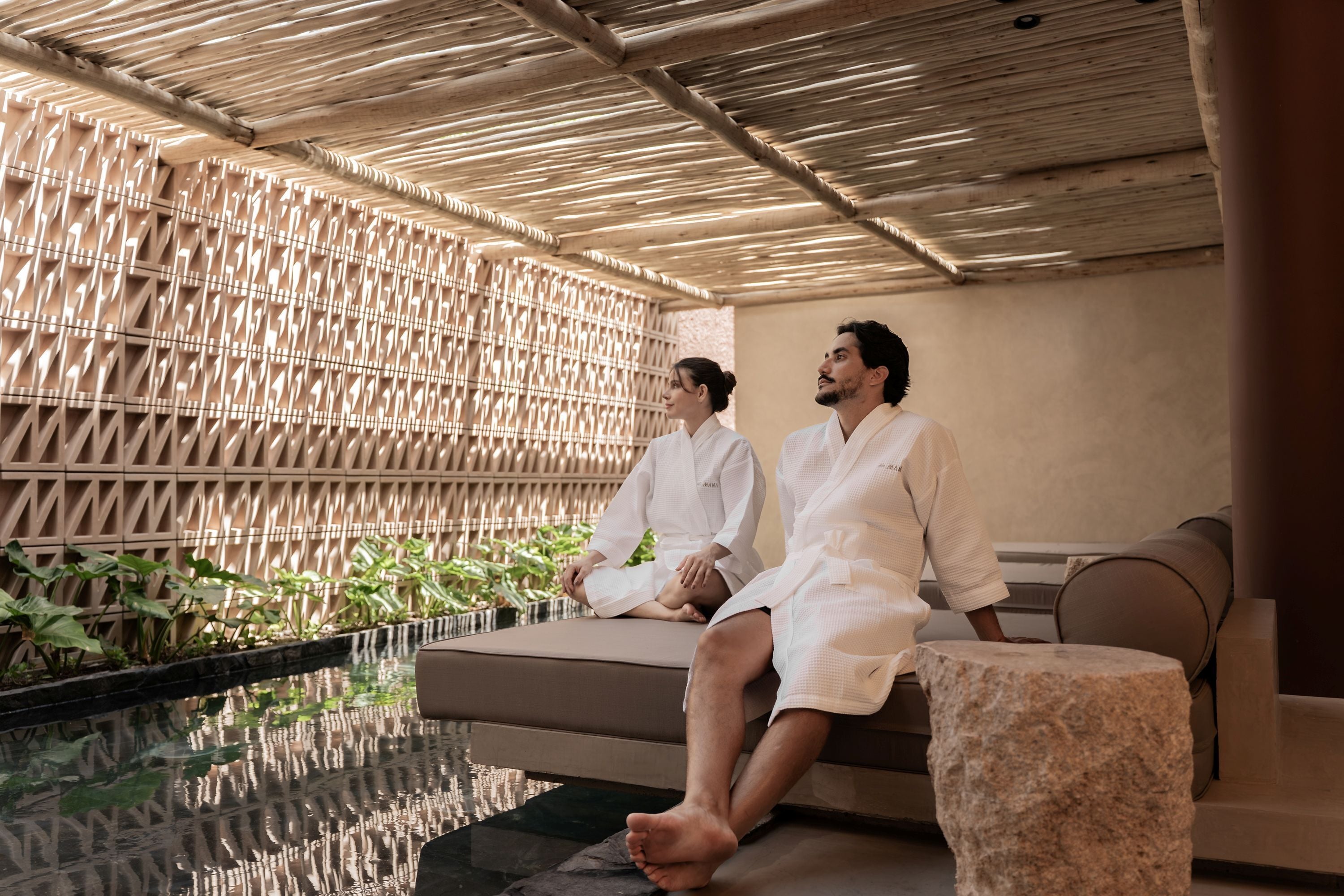 Two people in white robes sitting on a bench by a pool with a bamboo lattice wall and greenery in the background.