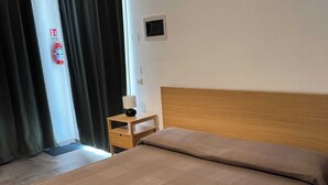A hotel room with a bed, a nightstand, a lamp, a fire extinguisher, and a door with curtains. - Tremiti (Vico del Gargano)