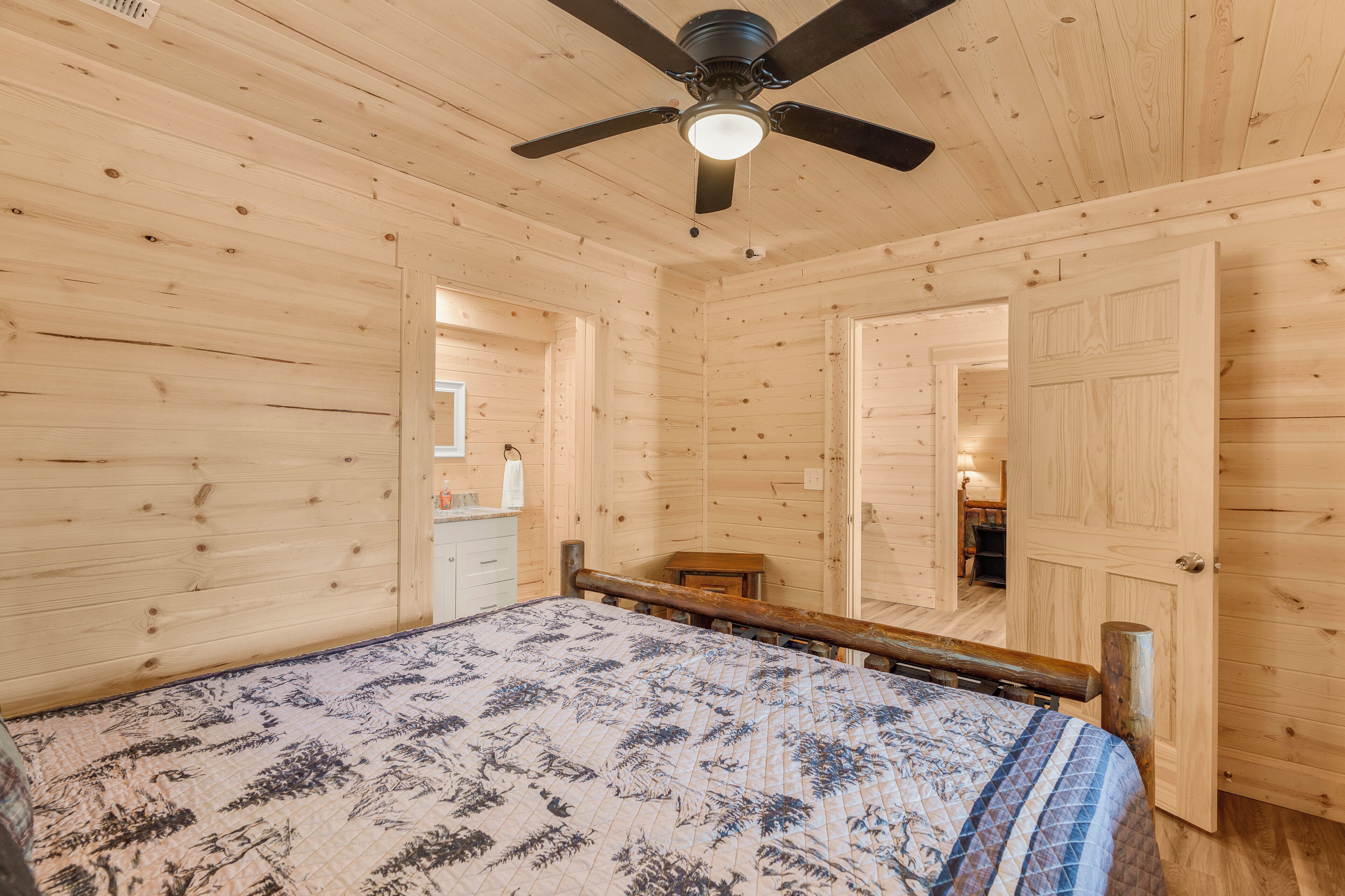 Un dormitorio de cabaña de madera con una cama, un ventilador de techo y una puerta que da a otra habitación.