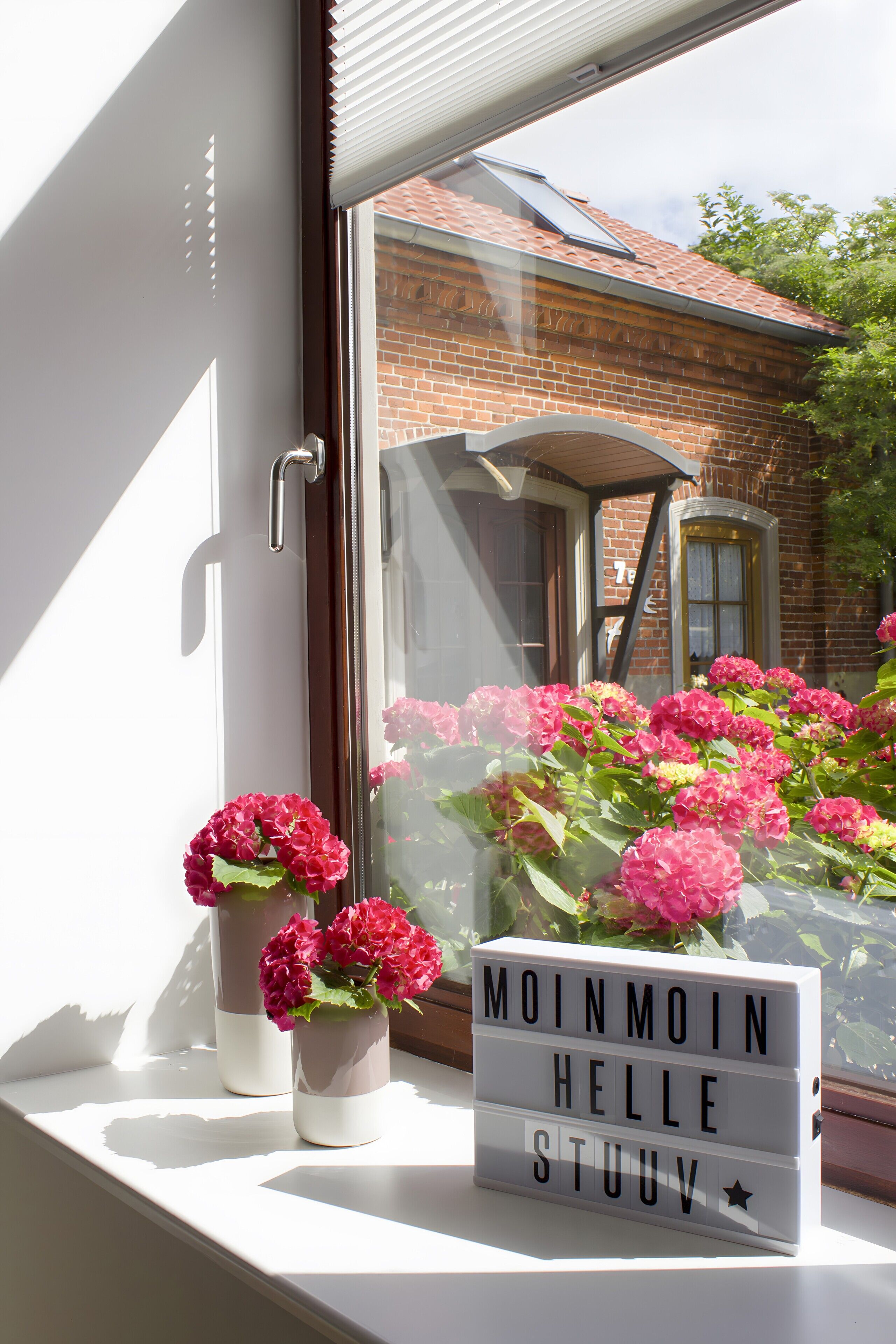 Una ventana con una caja de luz que muestra 'MOINMOIN HELLE STUUV' y dos jarrones con flores rosadas.