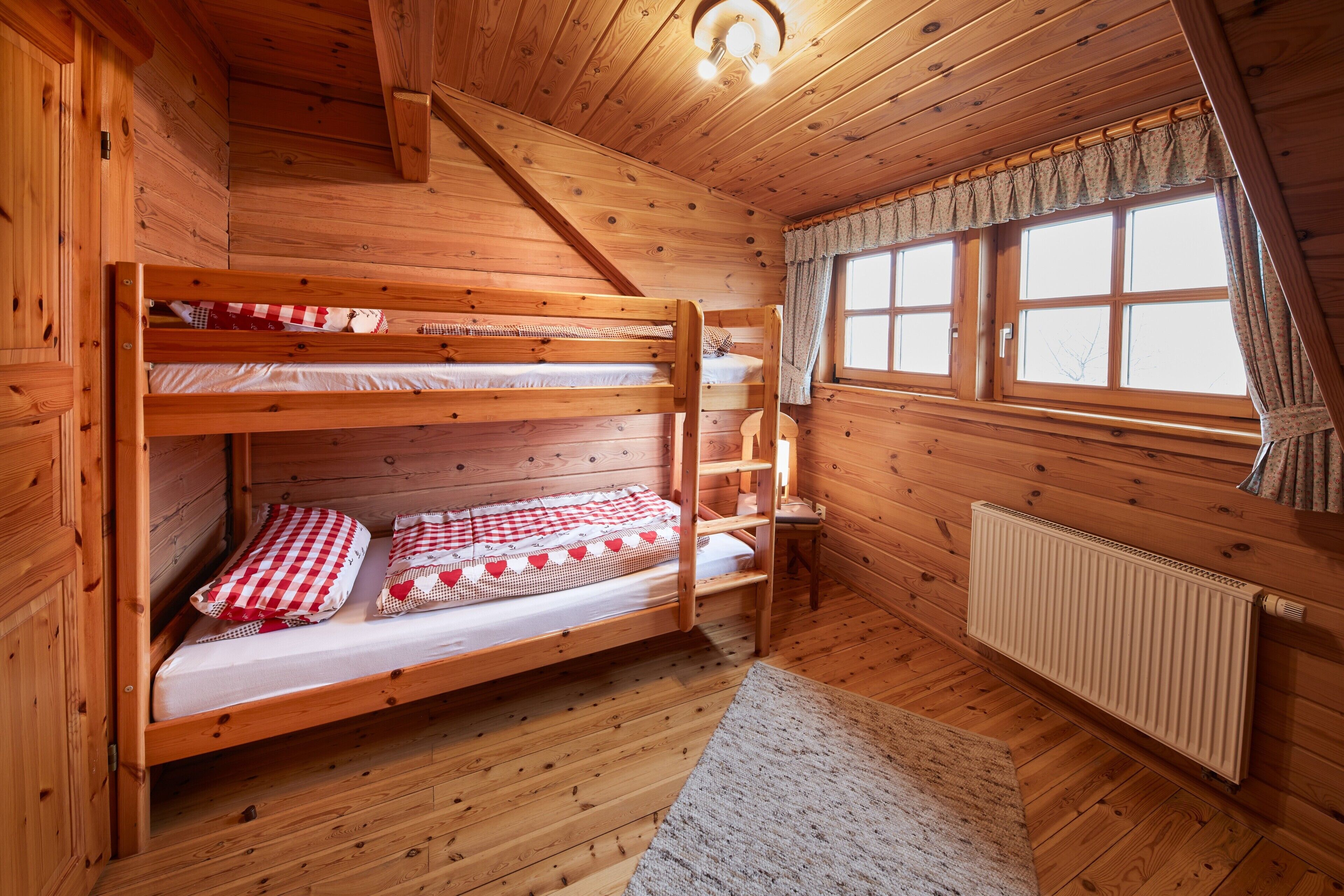 A wooden cabin room with bunk beds, a window with curtains, and a radiator.