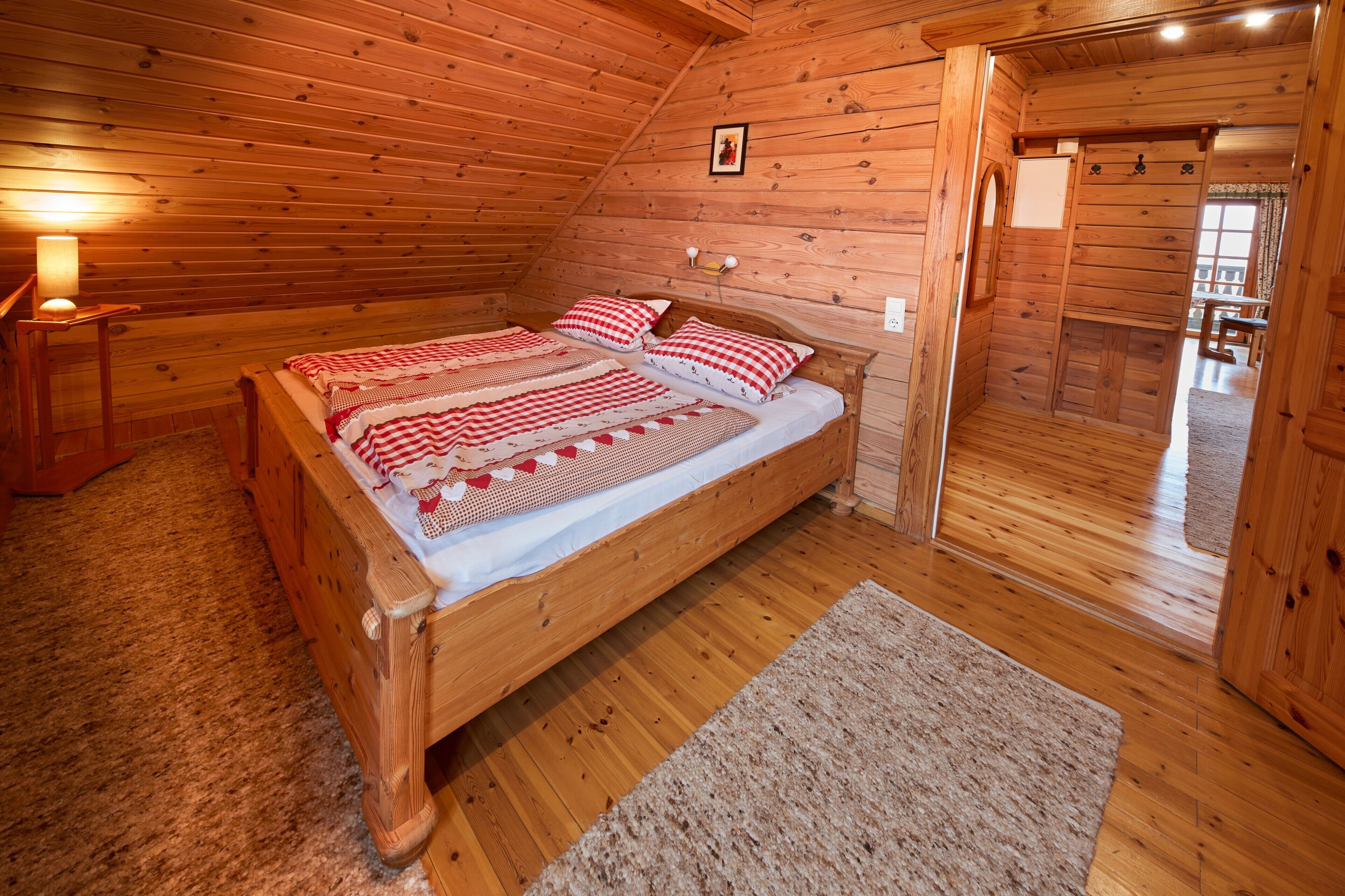 A wooden cabin bedroom with a bed, a nightstand, a lamp, and a view of the outside.