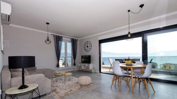 A modern living room with a sofa, a coffee table, and a dining area with chairs. There is a TV, a clock, and a large window offering a view of the sea. - Apartment Nina - panoramic sea views (Slatine)