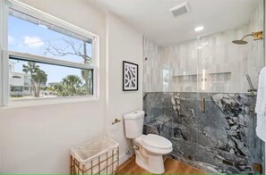 Un baño con inodoro, ducha, paredes de mármol, ventana y un cuadro en la pared.