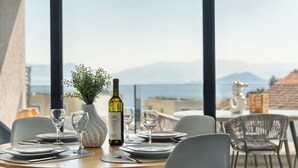 Mesa de comedor dispuesta con platos, cubiertos y copas de vino, junto con una botella y un florero con follaje. La mesa está colocada para disfrutar de vistas al mar y a las montañas al fondo.