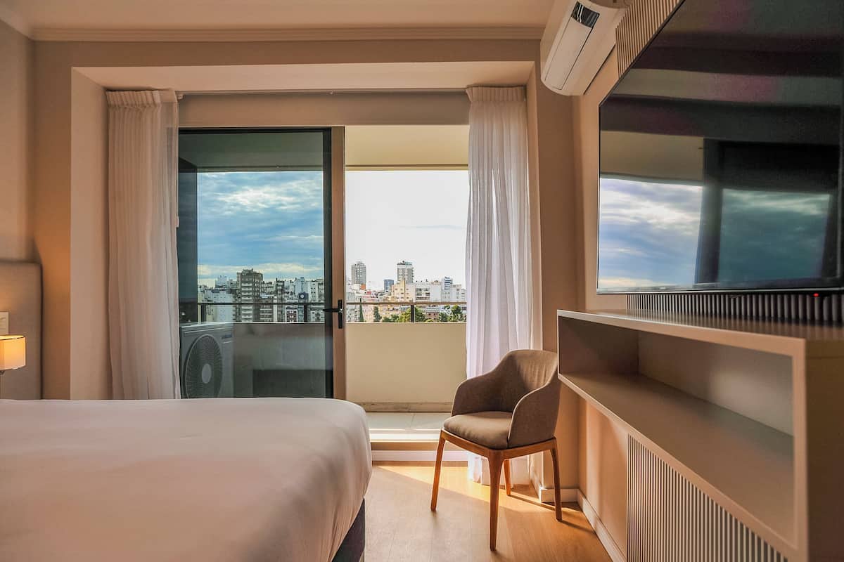 A hotel room with a bed, a chair, a TV, and a balcony with a city view.