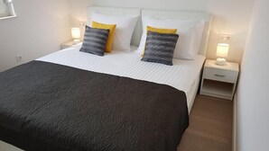 A neatly made bed with a dark patterned bedspread, two white pillows with a yellow accent, and a white headboard. There are two bedside tables with lamps, one on each side of the bed. - Vlads' Place - pool & garden-view (Slatine)