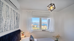 A bedroom with a bed, a nightstand, a vase with dried flowers, a window with curtains, and a view of buildings and the sea.