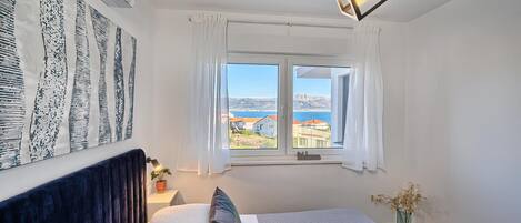 A bedroom with a bed, a nightstand, a vase with dried flowers, a window with curtains, and a view of buildings and the sea.
