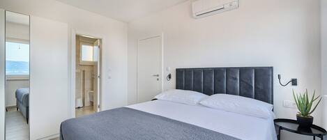 A bedroom with a bed, a bedside table with a plant, a mirror, and an air conditioning unit.