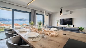 A modern dining area with a wooden table set for four, large windows offering a view of the sea and buildings, a flat-screen TV mounted on the wall, and a minimalist decor style. - Aqualis Vista (Slatine)