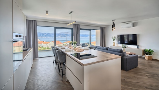 A modern kitchen with a central island, dining area, and a living space with a TV and plants. - Aqualis Vista (Slatine)