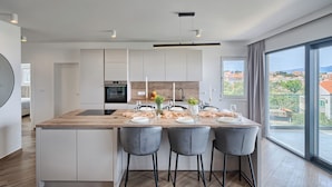 A modern kitchen with a central island, bar stools, and a view of the city through large windows. - Aqualis Vista (Slatine)