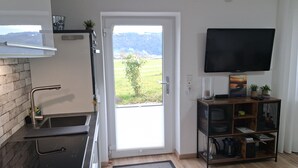 A modern kitchen with a sink, a stove, and a door leading to a balcony. There is a television mounted on the wall and a wooden shelf with various items. - Apartment 'Lounge' with Mountain View, Private Terrace and Wi-Fi (Sigmarszell)