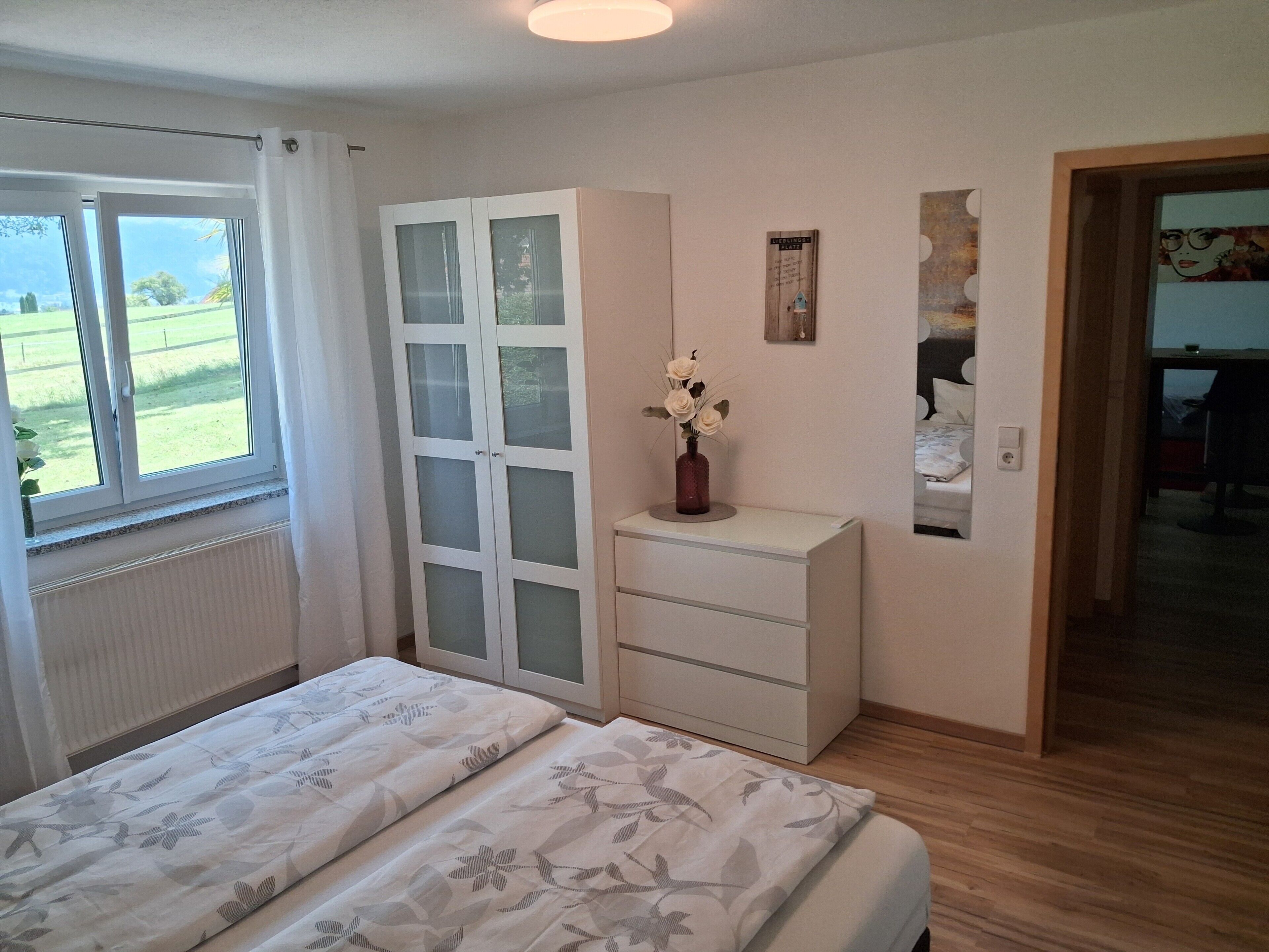 Un dormitorio con cama, armario, una mesita con un florero y vistas al exterior.