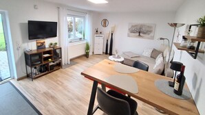 A modern living room with a wooden dining table, a sofa, a TV mounted on the wall, and a window with curtains. - Apartment 'Lounge' with Mountain View, Private Terrace and Wi-Fi (Sigmarszell)