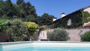 A swimming pool with a clear blue water surface, surrounded by a garden with various plants and trees, and a building with a tiled roof and ivy-covered walls. - Saint Privat House: Pool, Family & Greenery (Saint Privat en Périgord)
