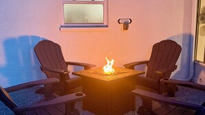 Une terrasse avec deux fauteuils Adirondack, un foyer carré avec une flamme bien visible, et une fenêtre donnant sur le ciel.