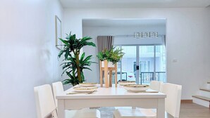 A modern dining area with a white table set for four, surrounded by white chairs. There is a potted plant on the table and another on the floor. The room has a wooden floor and white walls. - Minimalist Townhome Stay (Pattaya)