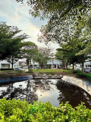 A serene pond with a metal bridge, surrounded by lush greenery and trees, with buildings in the background. - Minimalist Townhome Stay (Pattaya)