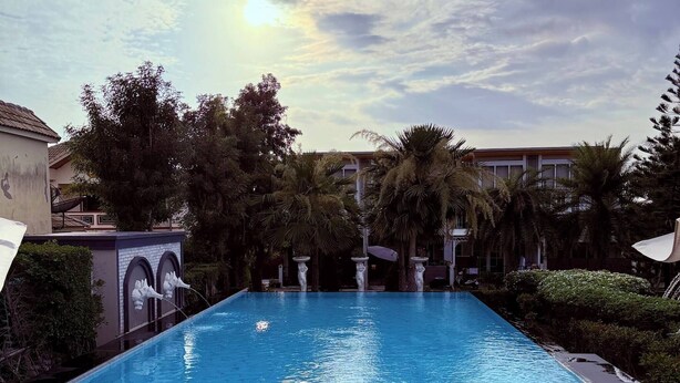 A rectangular swimming pool with clear blue water, surrounded by palm trees and a building with a tiled roof. - Minimalist Townhome Stay (Pattaya)