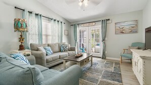 A living room with a sofa, a coffee table, and a TV stand. There is a door labeled "Beach House" and a window with curtains. - 3B/2B house with POOL. 700' to public boat ramp & .6 mi to beach access! (Panama City Beach)