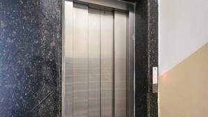 Un ascenseur à l’intérieur métallique, situé dans un couloir dont le mur est de couleur noire et beige, avec des motifs pointillés.