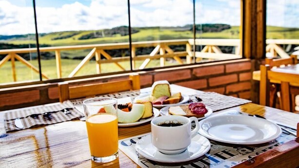 A breakfast table with a glass of orange juice, a cup of coffee, and various pastries, set against a scenic view through large windows. - Pousada Morada da Serra (Cambara do Sul)