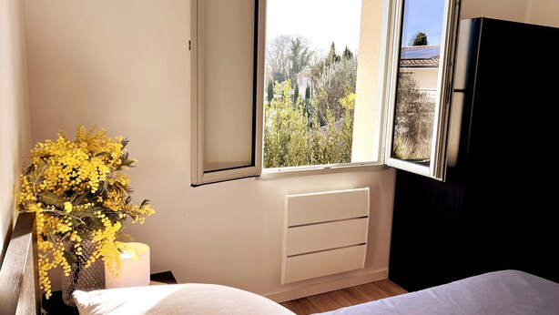 A bedroom with a window, a radiator, a bed, and a nightstand with a lamp and flowers. - Spacious House in Libourne – Near Saint-Émilion and Bordeaux (Libourne)