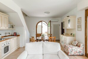 Uma sala ampla com um sofá branco, uma mesa de jantar com cadeiras e uma cozinha com armários brancos e uma chaleira vermelha.