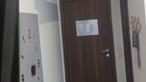 Ein Flur mit einer Holztür, einer Uhr und einem roten Schreibtisch mit Fernbedienung.