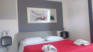 Una habitación de hotel con una cama, almohadas blancas, una colcha roja, una obra de arte enmarcada en la pared y un perchero con toallas.