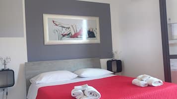 Ein Hotelzimmer mit einem Bett, weißen Kissen, einer roten Bettdecke, einem gerahmten Kunstwerk an der Wand und einem Handtuchhalter mit Handtüchern.