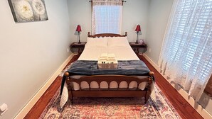 Un dormitorio con una cama de madera, dos mesitas de noche con lámparas, una ventana con cortinas de encaje y una alfombra estampada.