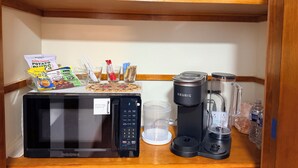 Una zona de cocina con microondas, cafetera Keurig y licuadora sobre la encimera, junto con un hervidor, tazas y una bolsa de patatas fritas.